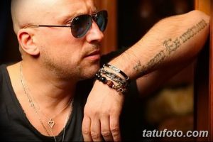Фото тату: мужчина в солнцезащитных очках с татуировкой на предплечье, выполненной готическим шрифтом. На руке несколько браслетов.