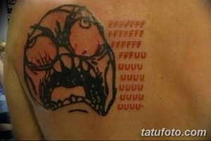 Фото тату с изображением популярного интернет-мема Rage Face, выполненного в черном цвете с красными надписями, на плече.