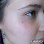Фото пирсинга брови с серебристым шариком на девушке с темными волосами и татуировкой на шее. Крупный план лица.