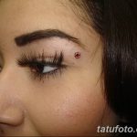 Фото пирсинга брови с красным камнем на девушке с темными волосами и выразительными глазами. Крупный план лица.