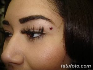 Фото пирсинга брови с красным камнем на девушке с темными волосами и выразительными глазами. Крупный план лица.