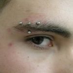 Фото тату: пирсинг брови с множеством серебристых проколов над бровью. Альтернативный стиль, акцент на глазах.