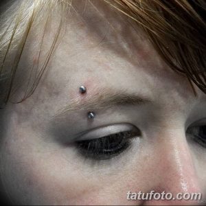 Фото пирсинга брови с двумя серебристыми украшениями. Крупный план женского лица с акцентом на глаза и пирсинг.
