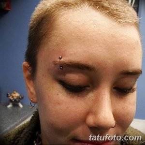 Фото пирсинга брови с двумя синими украшениями. Крупный план женского лица с акцентом на глаза и пирсинг.