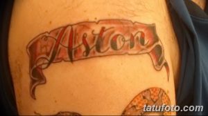 Фото тату надписи 'Aston' выполненной в стиле олдскул на ленте, расположенной на плече. Красные и черные цвета, четкие контуры.