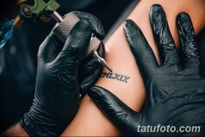 Фото тату: мастер татуировки в черных перчатках наносит татуировку римскими цифрами на кожу клиента. Видна тату-машинка.