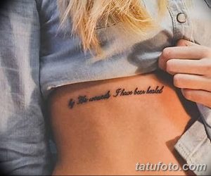 Фото тату с надписью "By the wounds I have been healed" на женском боку. Минималистичный дизайн и элегантный шрифт делают татуировку стильной и значимой.