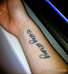 Фото тату с надписью "stay strong" и маленьким сердечком на запястье. Черно-белый рисунок татуировки, выполненный тонким шрифтом.