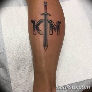 Фото татуировки на икре: меч, пронзающий инициалы 'K' и 'M', выполненные в готическом стиле.
