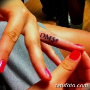 Фотография татуировки с надписью 'DMS' на пальце руки. Текст выполнен тонким шрифтом, цвет татуировки соответствует тону кожи.