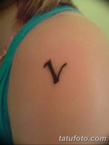 Фото татуировки на плече с изображением буквы 'V' в каллиграфическом стиле. Черно-белый рисунок, тонкие линии, простой и элегантный дизайн.