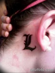 Фото тату: стильная татуировка с буквой 'L', расположенная за ухом. Черный рисунок татуировки на светлой коже, минималистичный дизайн.
