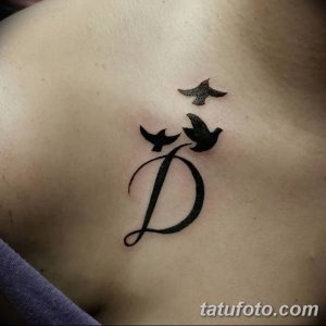 Фото тату: буква 'D' в каллиграфическом стиле с двумя летящими птицами над ней. Черно-белая татуировка на ключице.