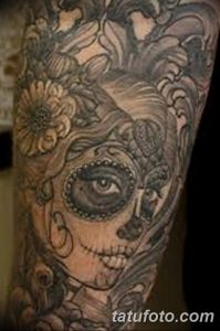 Фото татуировки на руке с изображением Sugar Skull, окруженного цветами и детальной штриховкой. Реалистичный черно-серый рисунок.