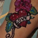 Фото тату с использованием блесток, изображающее розовую розу и надпись LOVE. Яркий и привлекательный рисунок для тех, кто любит выражать свои чувства.