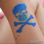 Фото тату с изображением черепа и костей, выполненного с использованием синих и зеленых блесток. Татуировка нанесена на предплечье.