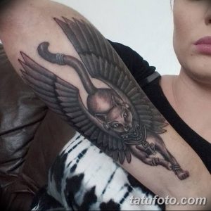 Фото тату египетской кошки с крыльями на руке. Детализированный рисунок с использованием серых и коричневых оттенков.