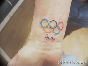 Фото тату: Цветная татуировка с изображением олимпийских колец и надписью 'Judo' на запястье. Символичный рисунок для спортсмена.