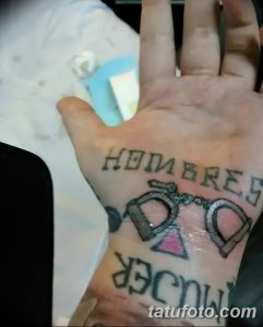 Фото тату на ладони с надписями и изображением пистолета. Черно-серый рисунок татуировки выполнен в стиле олдскул с элементами граффити.