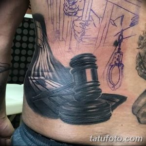 Фото реалистичной татуировки с изображением молотка правосудия и наручников на теле. Черно-серый рисунок татуировки с детальной проработкой теней.