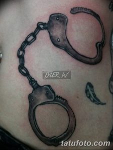 Фото реалистичной татуировки наручников с цепью на боку. Детализированный рисунок в черно-серых тонах.
