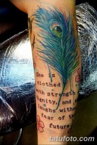 Фото тату на руке с изображением яркого пера павлина и цитаты 'She is clothed with strength and dignity...', выполненное в акварельном стиле.