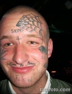 Фото мужчины с экстремальными татуировками на лице в стиле Skinhead. Изображены надписи, символы и рисунки, покрывающие большую часть лица.