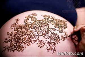 Фото тату мехенди на животе беременной женщины, с детально проработанными цветочными узорами в коричневых тонах. Мастер наносит рисунок.