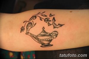 Фото тату лампы джинна, обвитой ветвью с листьями, на предплечье. Изящный рисунок в минималистичном стиле с тонкими линиями и деталями.