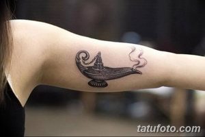 Фото тату: рисунок лампы Алладина с клубами дыма, выходящими из носика, на руке, выполненный в черно-серых тонах.