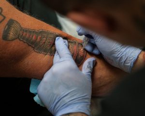 Фотография, демонстрирующая процесс нанесения татуировки на руку мужчины. Мастер в перчатках работает над татуировкой, используя тату-машинку.