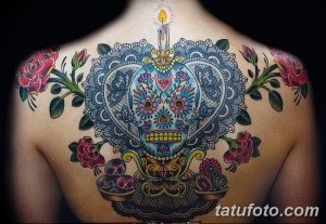 Фото тату: красочный рисунок сахарного черепа с цветами и свечой на спине. Детализированная татуировка в стиле Day of the Dead.