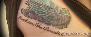Фото тату: цветная татуировка совы в полете с надписью "Nevertheless, She Persisted" на плече девушки. Вдохновляющий рисунок татуировки с сильным посланием.