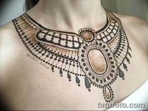 Фото тату хной на груди и шее женщины, имитирующее ожерелье с крупным кулоном. Выполнено в коричневых и черных тонах.