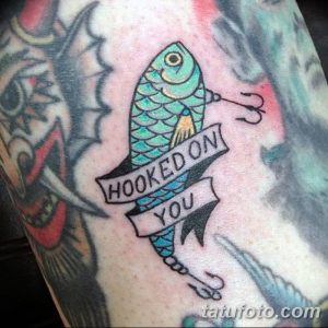 Фото тату с изображением яркой рыбы, пронзенной крючком, и надписью "Hooked On You". Цветной рисунок татуировки на руке.