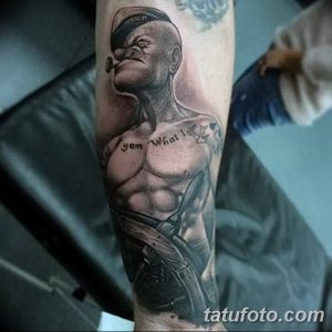 Черно-серая татуировка на руке с изображением мультяшного персонажа Попая-моряка, держащего штурвал. Рисунок выполнен в реалистичном стиле.