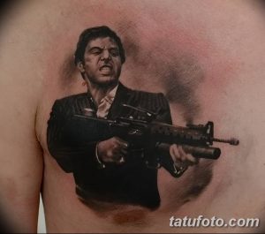 Фото тату реалистичного портрета Тони Монтаны из фильма «Лицо со шрамом» с автоматом в руках на груди мужчины. Детализированный черно-серый рисунок татуировки.