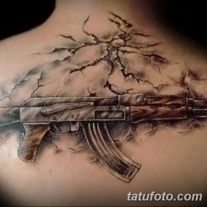 Фото тату реалистичного автомата Калашникова на спине, окруженного трещинами и молниями. Детализированный рисунок татуировки, выполненный в черно-серых тонах.