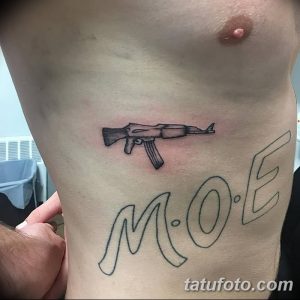 Фото тату небольшого автомата и надписи "MOE" на боку. Черно-серый контурный рисунок татуировки с минималистичным дизайном.