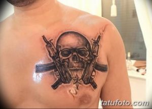 Фото тату черепа в очках, держащего два автомата. Черно-серая татуировка с реалистичной проработкой деталей и мрачной атмосферой.