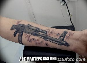 Фото тату реалистичного автомата Калашникова на предплечье, с имитацией повреждений и брызг краски, выполненное в серо-черных тонах.