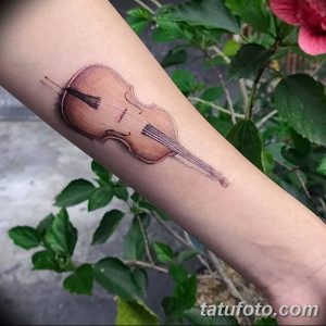 Фото реалистичной татуировки скрипки на предплечье. Изображение выполнено в коричневых и черных тонах с детальной проработкой текстуры дерева.