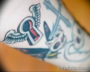 Татуировка на руке с изображением ключа, украшенного крыльями, выполненная в черно-серых тонах.