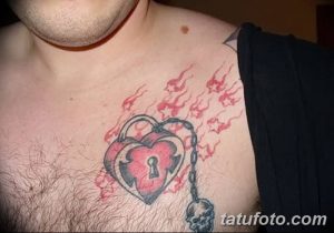 Фото тату на груди: изображение замка в форме сердца с открытым замочным отверстием, украшенного черепом и огненными брызгами. Драматичный и детализированный рисунок татуировки.