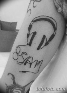 Фото татуировки на руке: наушники и имя "SAM", выполненные в черно-белом стиле, с элементами каллиграфии.