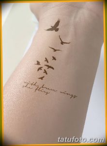 Фото тату на руке: стая силуэтов птиц, летящих вверх, и надпись 'with brave wings we fly', выполненные в коричневых тонах.