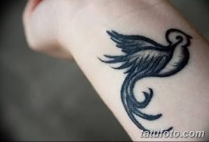 Фото татуировки с изображением стилизованной птицы на запястье. Тату выполнено в черном цвете с использованием плавных линий и завитков.