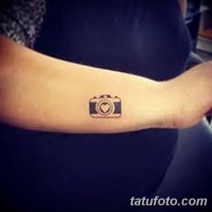 Фотография черно-белой татуировки на руке, изображающей фотоаппарат с сердцем вместо объектива. Простой и лаконичный рисунок тату.
