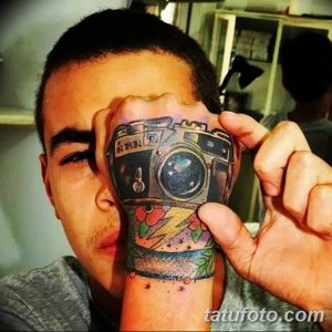 Фотография цветной татуировки на руке, изображающей фотоаппарат, как будто выглядывающий из кожи. Оригинальный и яркий рисунок тату.