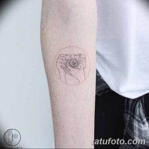 Фотография черно-белой татуировки на руке, изображающей руку, держащую старинный фотоаппарат. Минималистичный рисунок тату с тонкими линиями.